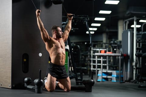 Serious attractive sportsman doing exercise for biceps, standing on knees in gym pulling cables