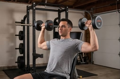 Man performing a shoulder press exercise with dumbbells in a home gym setting