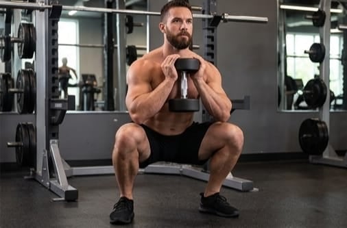 Man performing a dumbbell goblet squat exercise in a gym with mirrors and weight equipment visible in background